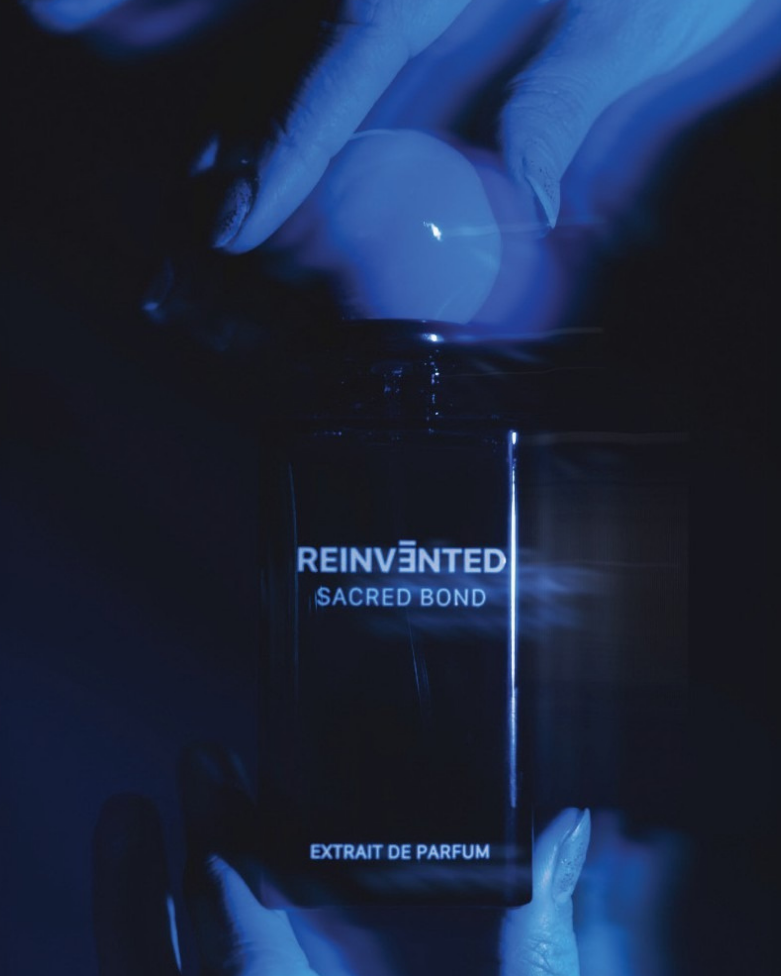 Image of Black perfume bottle labeled 'Reinvented Sacred Bond' held by a hand in dark lighting.
