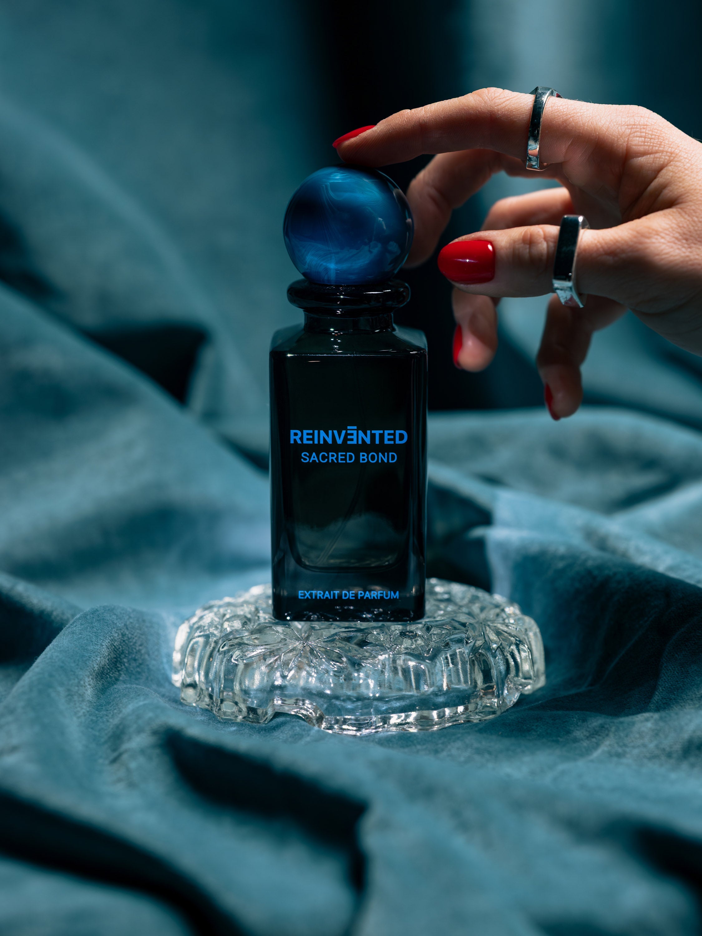 Image of Hand holding a blue perfume bottle labeled 'Reinvented Sacred Bond' on a textured blue fabric background